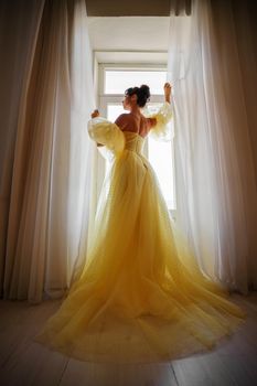 A woman's silhouette in a golden luxurious dress against the background of a window holds a curtain with her hands. Elegant lady in a yellow long silk dress with bare back, back view