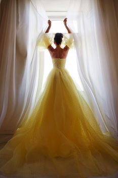 A woman's silhouette in a golden luxurious dress against the background of a window holds a curtain with her hands. Elegant lady in a yellow long silk dress with bare back, back view