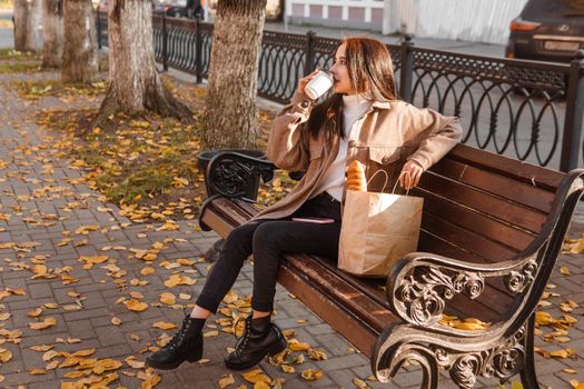 A stylish brunette woman walks around the autumn city. The brunette is sitting on a bench outside with a glass of coffee and a phone, holding a craft bag with a baguette.