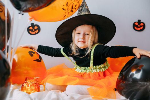 Children's Halloween - a girl in a witch hat and a carnival costume with airy orange and black balloons at home. Ready to celebrate Halloween.