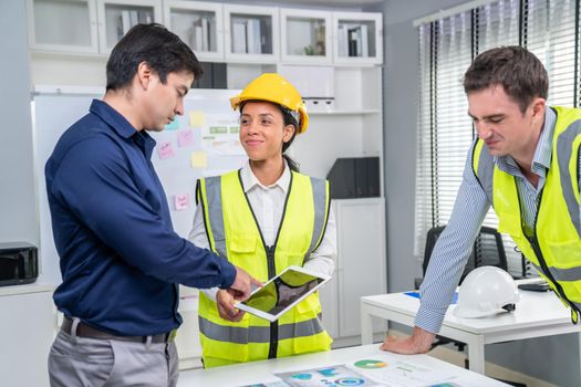 A team of competent engineers wearing safety equipment is working on blueprints with a tablet while also discussing with the investor in the office.