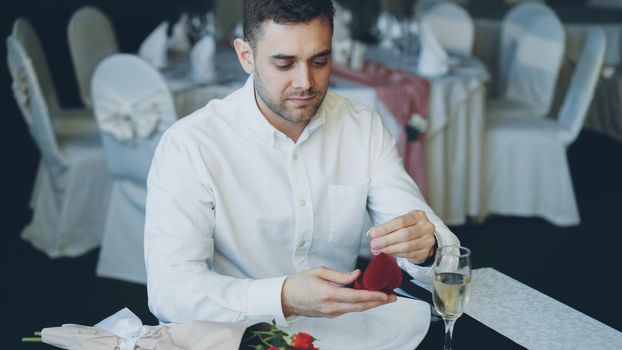 Loving handsome boyfriend is waiting for his girl in restaurant opening jewelry box and looking at ring. Bouquet of roses and glass of champagne are visible.