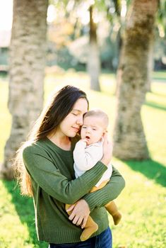 Mom hugs a sleepy baby in her arms, standing near palm trees. High quality photo