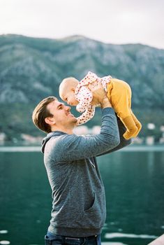 Smiling dad raises baby above his head against the background of water. High quality photo