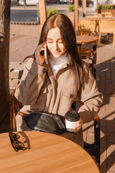 A stylish brunette woman is sitting at a table in an outdoor cafe with coffee and a phone. Autumn walk.