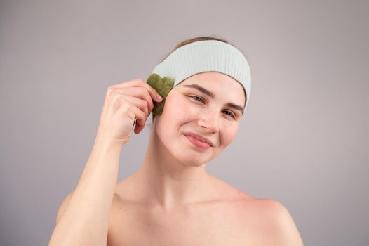 Portrait of a young woman massages her face with a gouache scraper on a white background