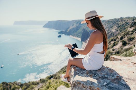 Successful business woman in yellow hat working on laptop by the sea. Pretty lady typing on computer at summer day outdoors. Freelance, travel and holidays concept.