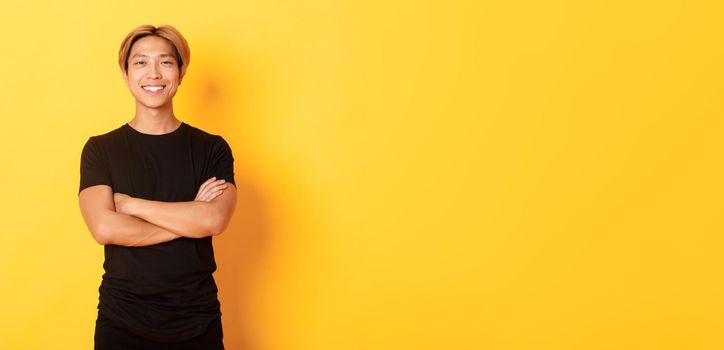 Portrait of confident handsome asian man smiling satisfied, standing over yellow background in black clothes.