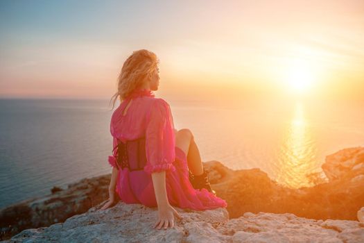 Side view a Young beautiful sensual woman in a red long dress posing on a volcanic rock high above the sea during sunset. Girl on the nature on blue sky background. Fashion photo