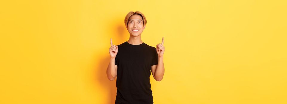 Handsome smiling asian man in black t-shirt pointing fingers up, looking at banner pleased, yellow background.