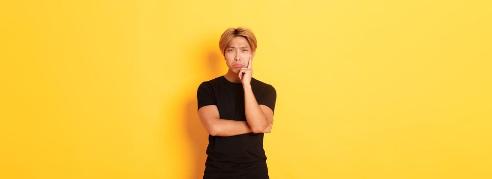 Upset thoughtful asian guy sulking and looking disappointed after thinking, standing yellow background.