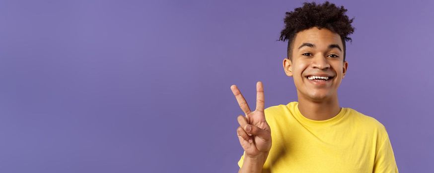 Close-up portrait of handsome upbeat young teenage guy with afro hairstyle, show peace sign and smiling, wear yellow t-shirt, staying optimistic and positive, purple background.