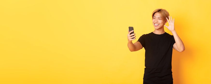 Portrait of friendly handsome asian man, smiling and waving hand at smartphone camera, greeting friends on video call, standing yellow background.