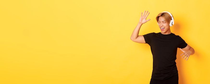 Happy attractive asian guy with blond hair, singing along and dancing as listening to music in wireless headphones, standing yellow background.