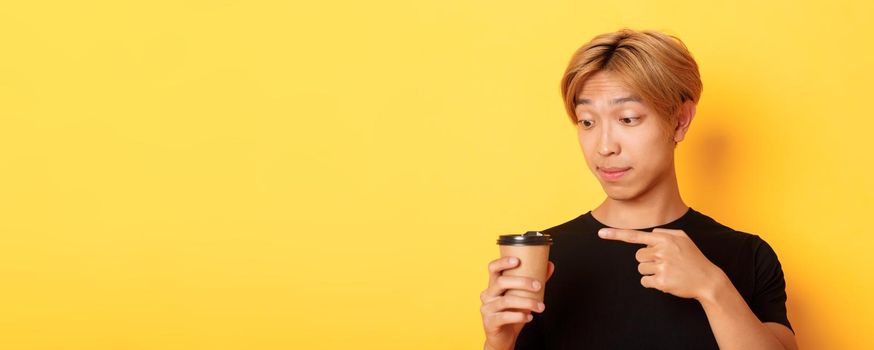 Close-up of impressed handsome asian guy pointing finger at delicious coffee, standing amazed over yellow background.