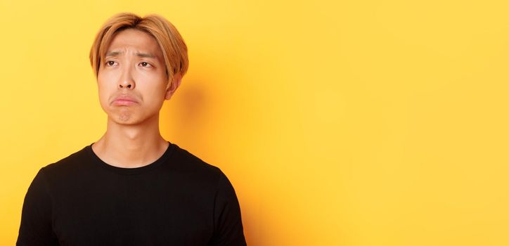 Close-up of sad and disappointed handsome asian guy sulking upset, looking upper left corner with regret or jealousy, standing yellow background.