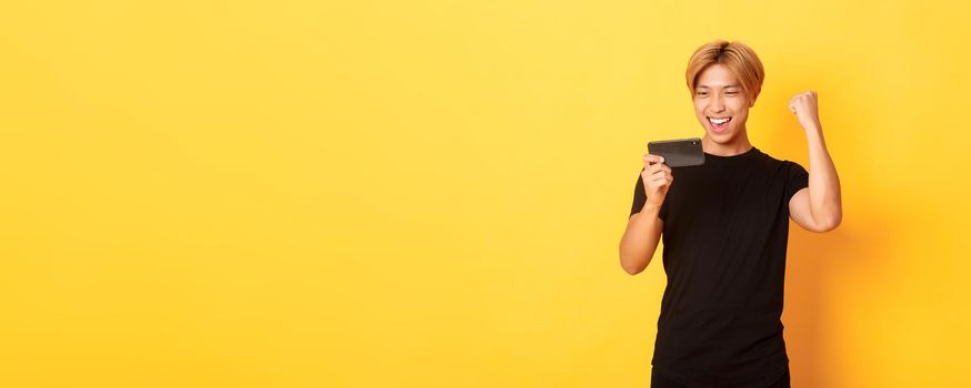Portrait of stylish handsome asian guy playing smartphone game, looking amused at mobile screen and rejoicing from winning, standing yellow background.