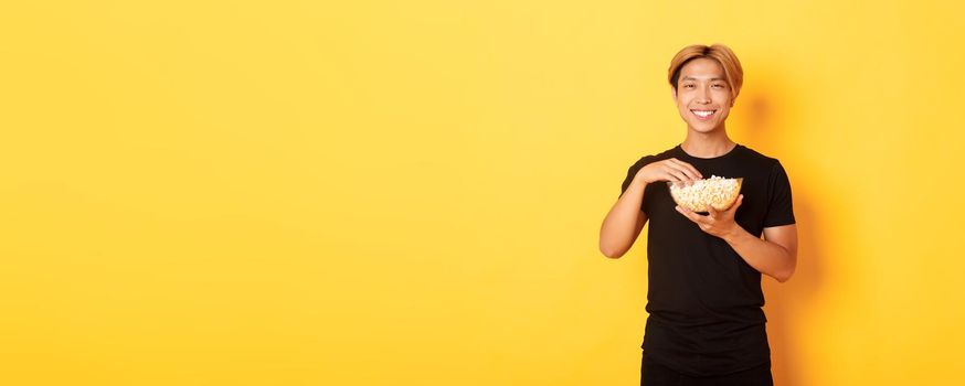 Pleased handsome korean guy smiling happy as enjoying watching movie or tv series, eating popcorn, standing yellow background.