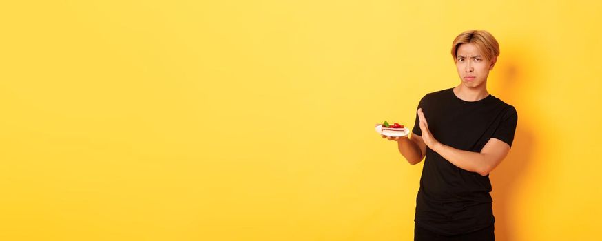 Portrait of disgusted and upset young asian guy refuse eating cake, looking displeased and showing rejection gesture, yellow background.
