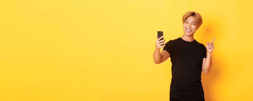 Portrait of handsome smiling asian man having video call on smartphone and pointing finger at something, standing yellow background.