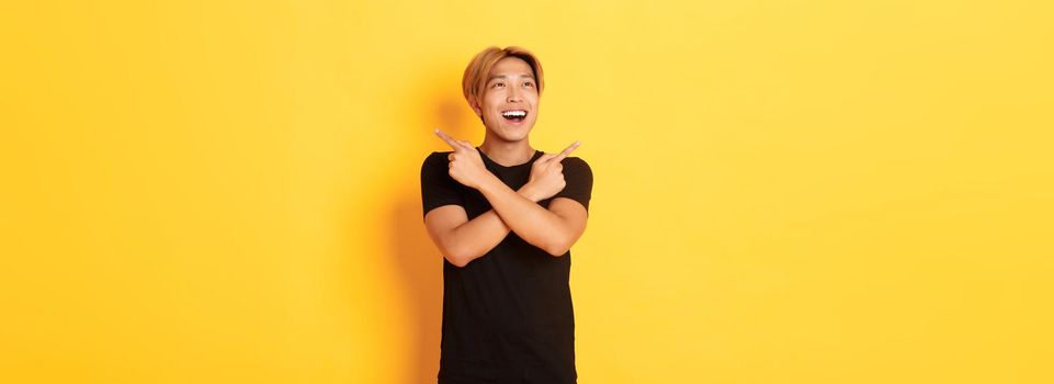 Portrait of happy smiling asian man with blond hair, looking amused left, pointing sideways at two variants, yellow background.