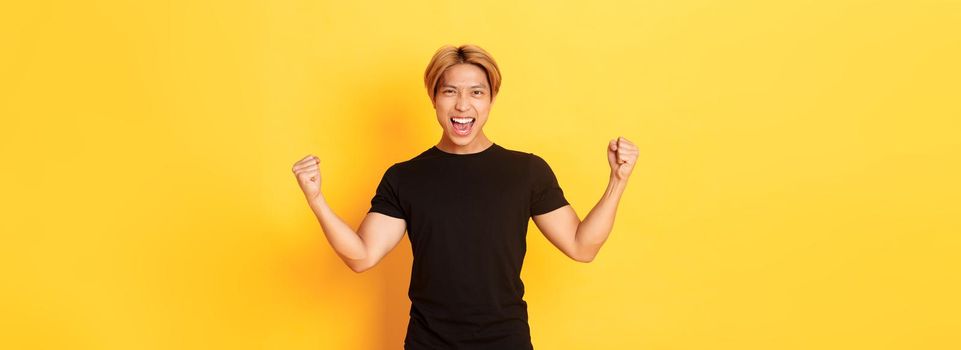 Portrait of happy attractive asian man rejoicing, raising hands up and shouting yes, standing pleased over yellow background.