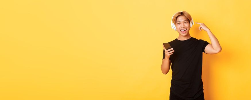 Happy satisfied asian guy likes music or podcast, pointing finger at headphones, holding smartphone, standing yellow background.