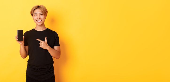 Portrait of handsome asian man showing something on smartphone screen, pointing at mobile phone display, standing over yellow background.