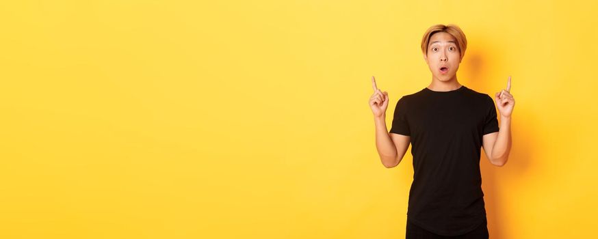 Portrait of astonished asian guy in black t-shirt, open mouth fascinated, pointing fingers up, yellow background.