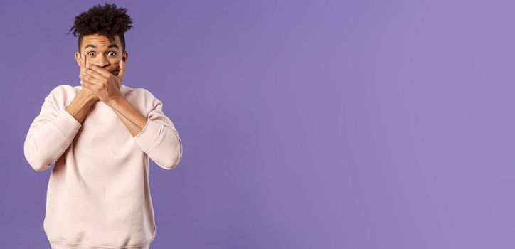 Portrait of shocked impressed young hispanic man found out gossip, heard rumour, trying not laugh shut his mouth with hands and express amazement and surprised with gaze, purple background.