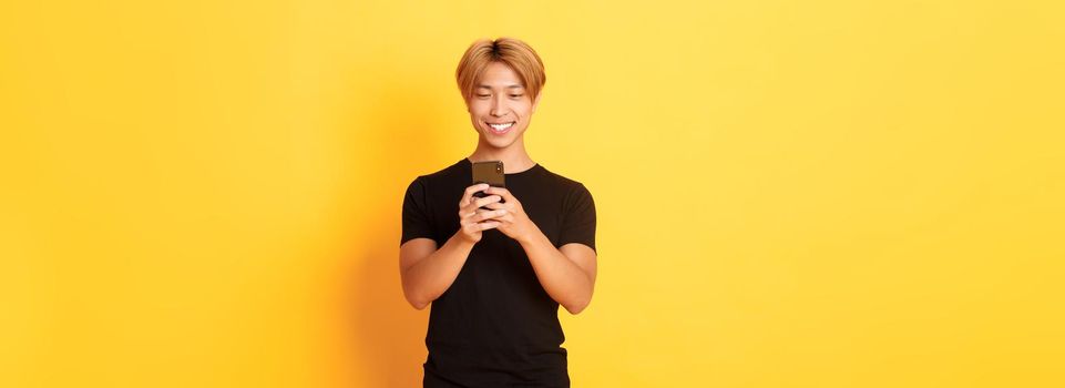 Portrait of handsome stylish asian guy with blond hair, using mobile phone and smiling, yellow background.