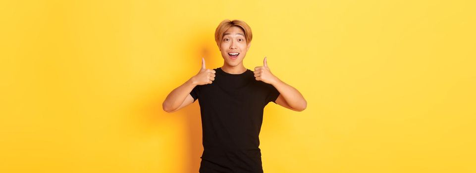 Portrait of amazed and pleased asian guy in black outfit, showing thumbs-up in approval, standing excited over yellow background.