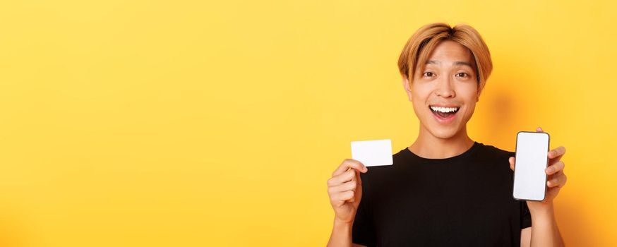 Amazed happy asian guy showing credit card and smartphone screen, smiling fascinated, standing yellow background.