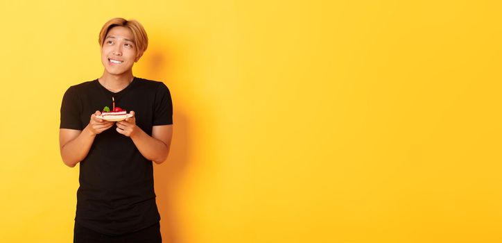 Portrait of handsome dreamy asian guy looking upper left corner and thinking, making wish while celebrating birthday and holding b-day cake, yellow background.