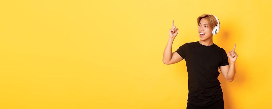 Joyful handsome asian guy with blond hair, singing along and dancing as listening to music in wireless headphones, standing yellow background.