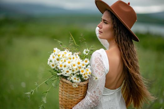 A middle-aged woman in a white dress and brown hat stands on a green field and holds a basket in her hands with a large bouquet of daisies. In the background there are mountains and a lake