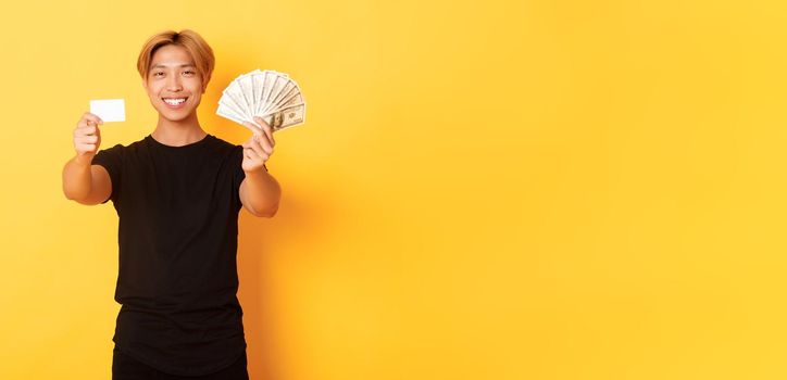 Satisfied attractive korean guy with blond hair, showing money and credit card, smiling happy, yellow background.