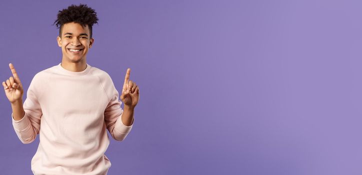 Waist-up portrait of joyful good-looking hispanic man with dreads, promoting product or company banner hanging on top, pointing fingers up, smiling satisfied, recommend subscribe or click link.