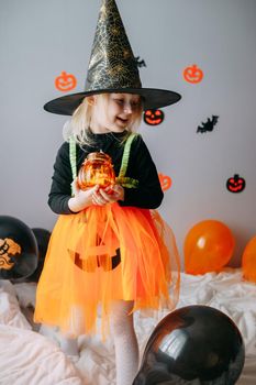 Children's Halloween - a girl in a witch hat and a carnival costume with airy orange and black balloons at home. Ready to celebrate Halloween.