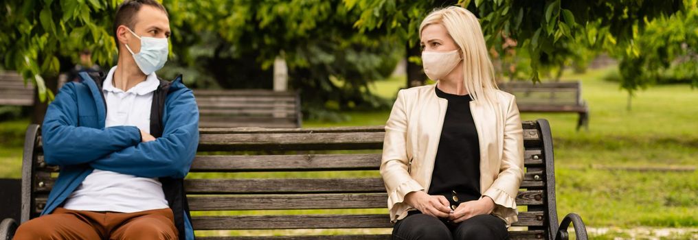 People sitting on park bench in the sun practicing social distancing in corona crisis wearing masks.