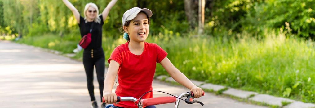 Beautiful young mother teaching her daughter to ride a bicycle. Both smiling, summer park in background, active family concept.