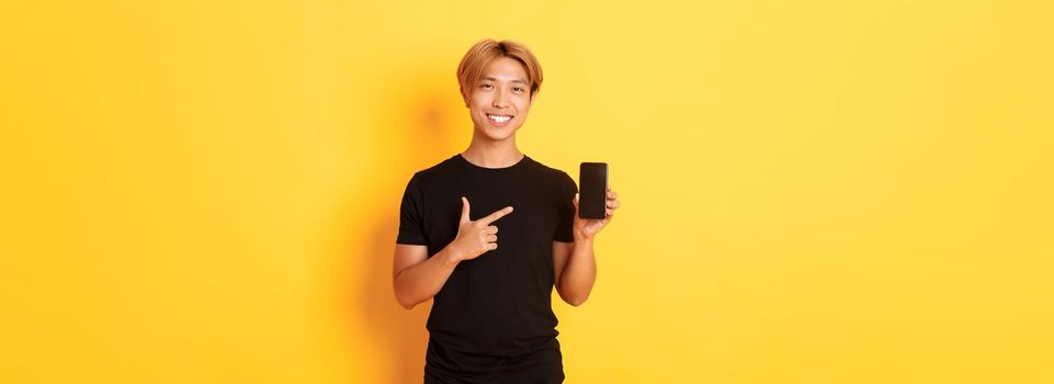 Portrait of handsome asian man showing something on smartphone screen, pointing at mobile phone display, standing over yellow background.