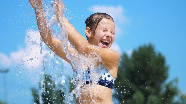 smiling, happy eight year old girl in swimsuit having fun in splashes in street city fountain, outdoors, in park, summer, hot sunny day during vacation. High quality photo