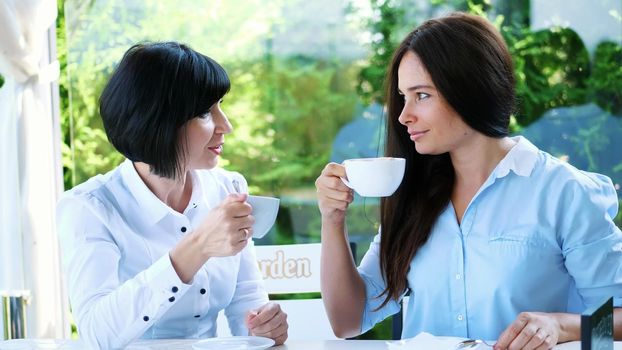 in the summer, on a terrace in a cafe, two forty-year-old beautiful brunettes of a woman are chatting cute, they are dressed in blond blouses. High quality photo