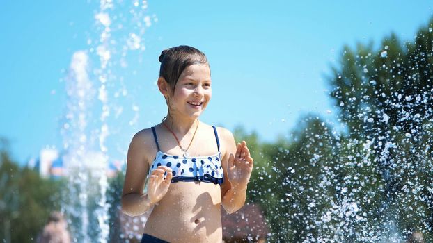 smiling, happy eight year old girl in swimsuit having fun in splashes in street city fountain, outdoors, in park, summer, hot sunny day during vacation. High quality photo