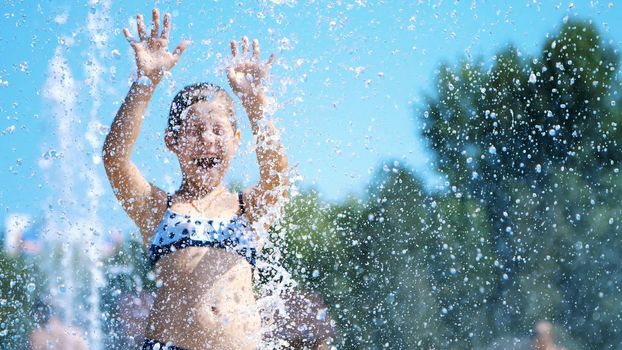 smiling, happy eight year old girl in swimsuit having fun in splashes in street city fountain, outdoors, in park, summer, hot sunny day during vacation. High quality photo