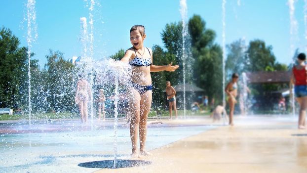 smiling, happy eight year old girl in swimsuit having fun in splashes in street city fountain, outdoors, in park, summer, hot sunny day during vacation. High quality photo