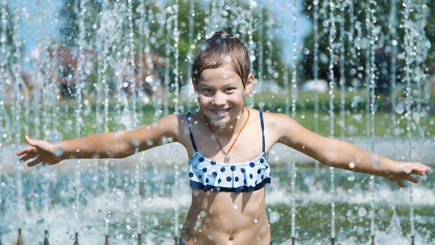 smiling, happy eight year old girl in swimsuit having fun in splashes in street city fountain, outdoors, in park, summer, hot sunny day during vacation. High quality photo