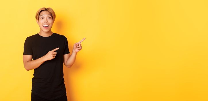 Amazed handsome korean guy, smiling fascinated and pointing fingers right, showing way, standing yellow background.