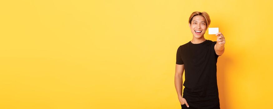 Cheerful handsome asian guy smiling pleased and showing credit card, standing yellow background.